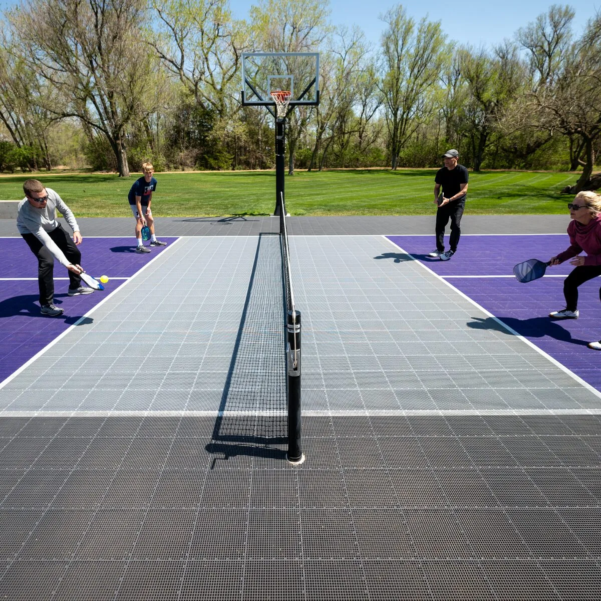 A group of four play pickleball on an outdoor pickleball court featuring a basketball hoop